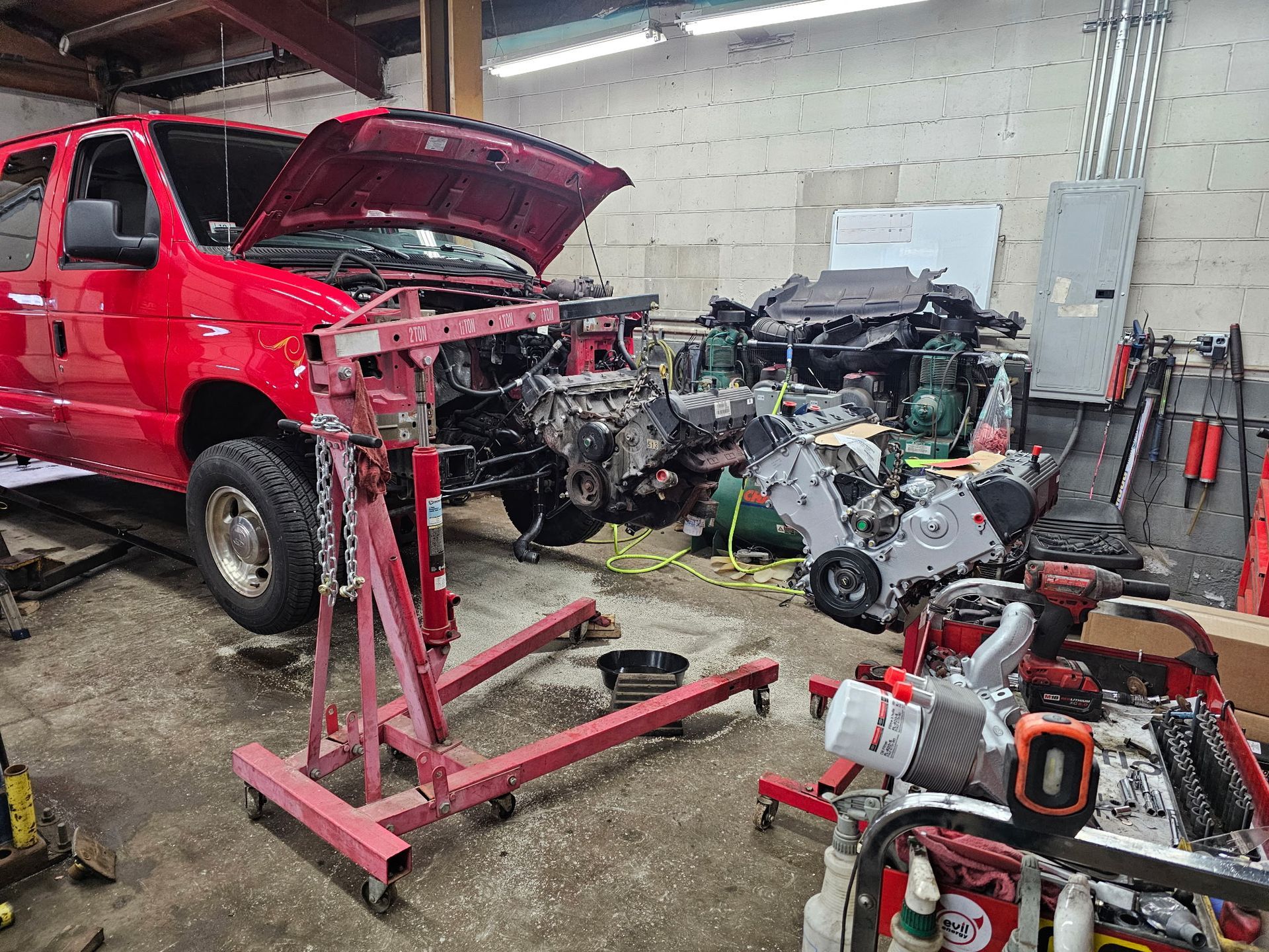 Red truck in an auto shop with hood open, surrounded by vehicle parts and repair equipment.