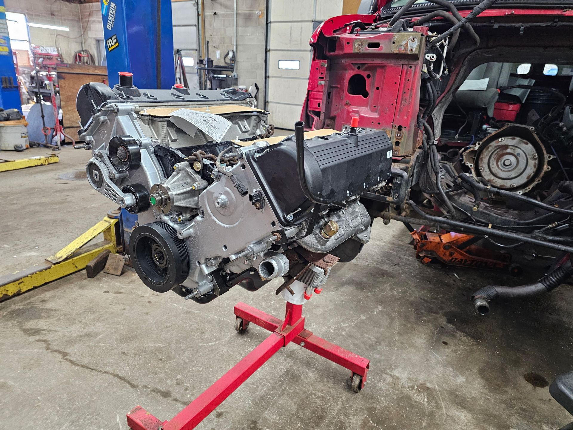 Car engine on a red stand inside a garage, with a partly disassembled vehicle behind it.