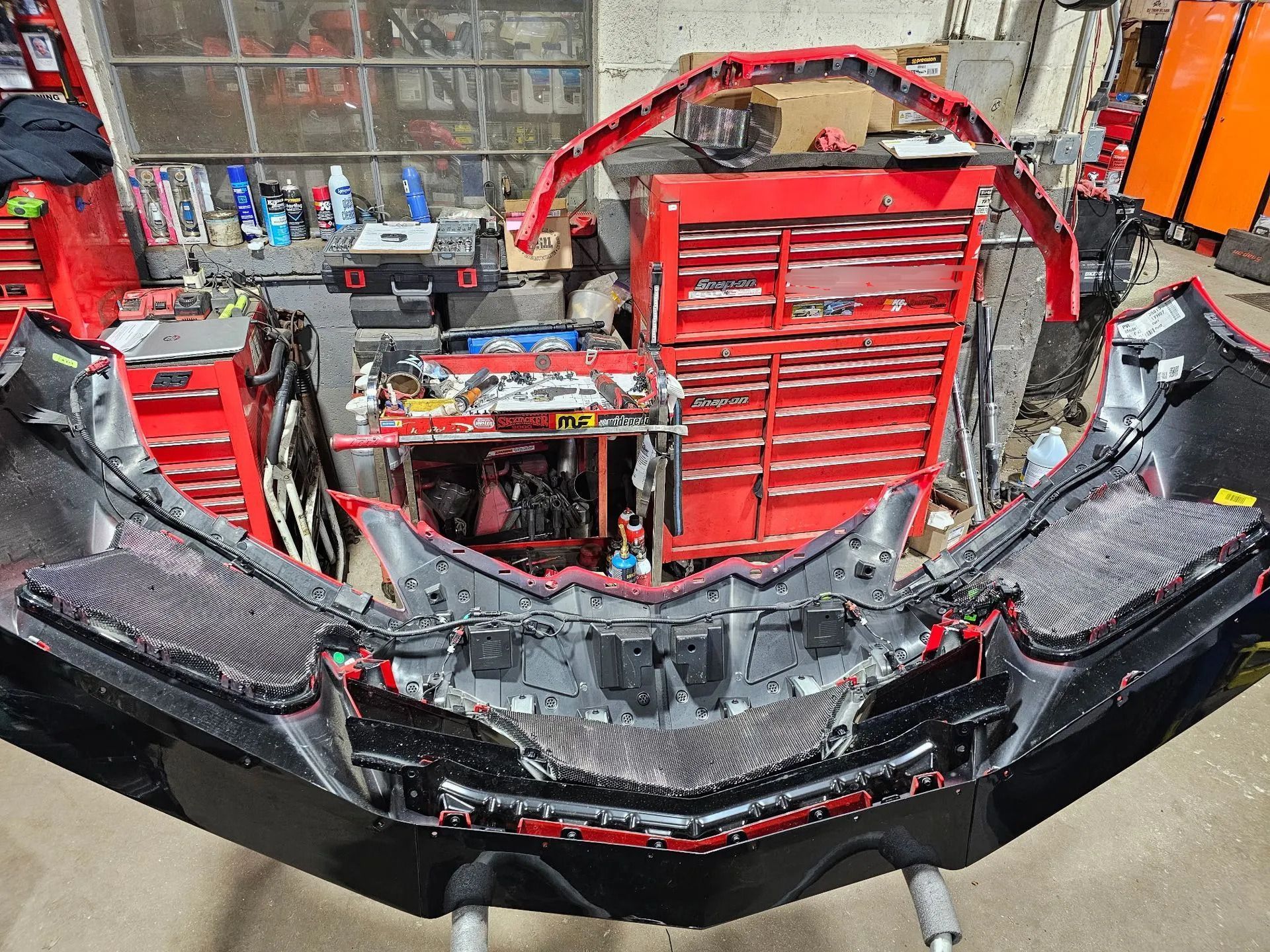 Front end of a red race car in a garage, with the bumper removed and internal components exposed.
