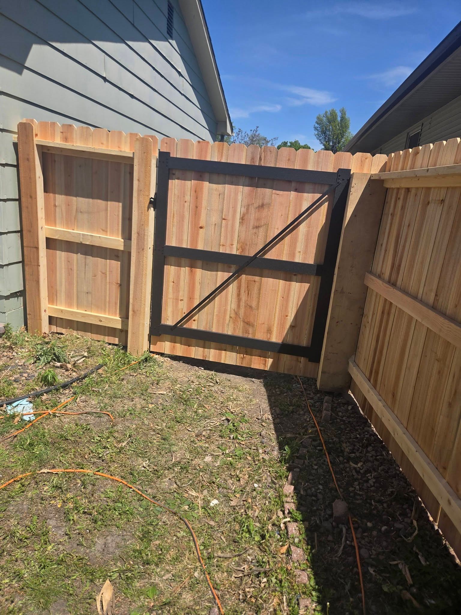 Wooden backyard gate with black cross brace between fences, beside a house and grassy yard.
