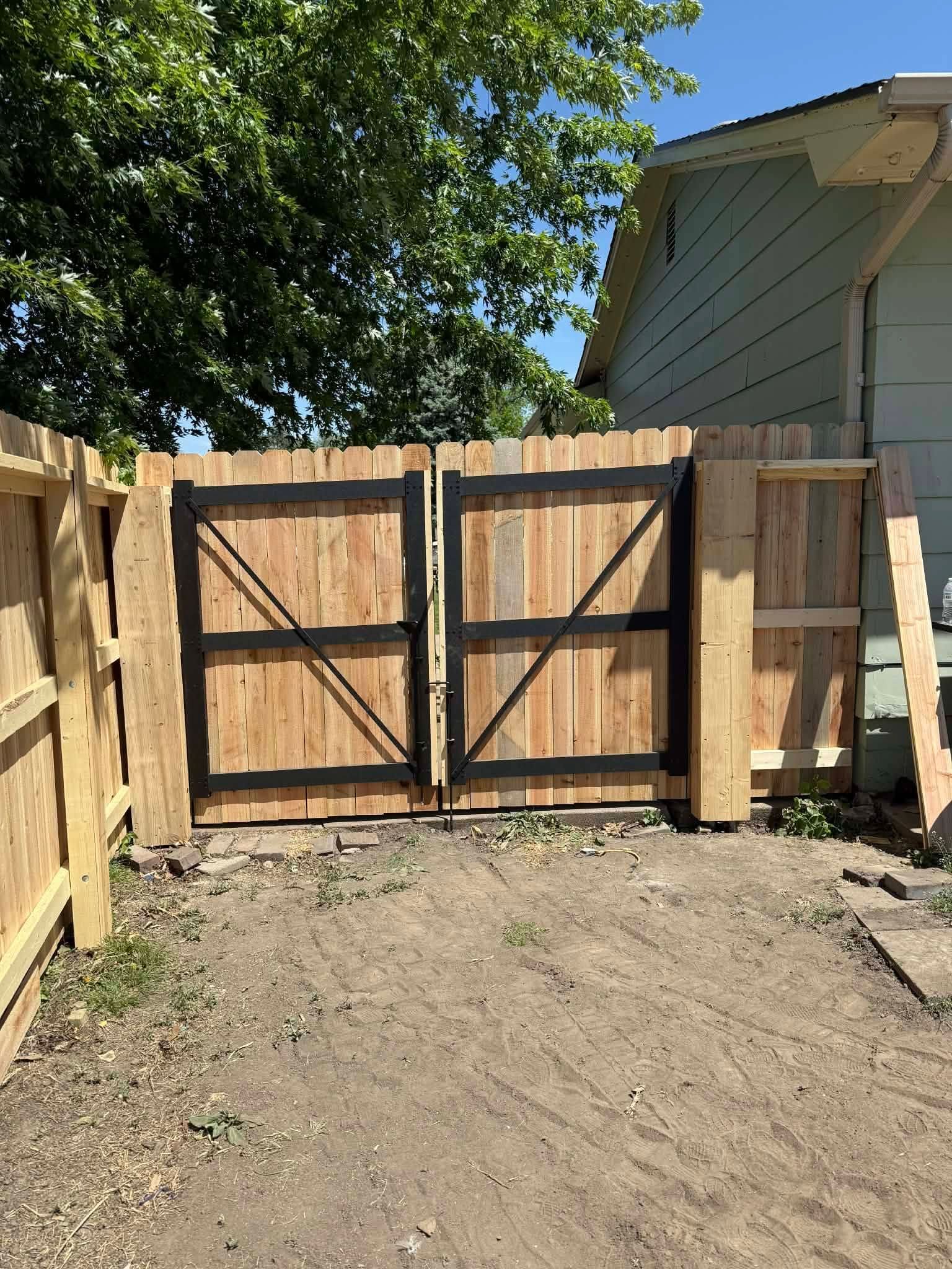 Wooden double gate between fences in a dirt backyard, with trees and a house visible.