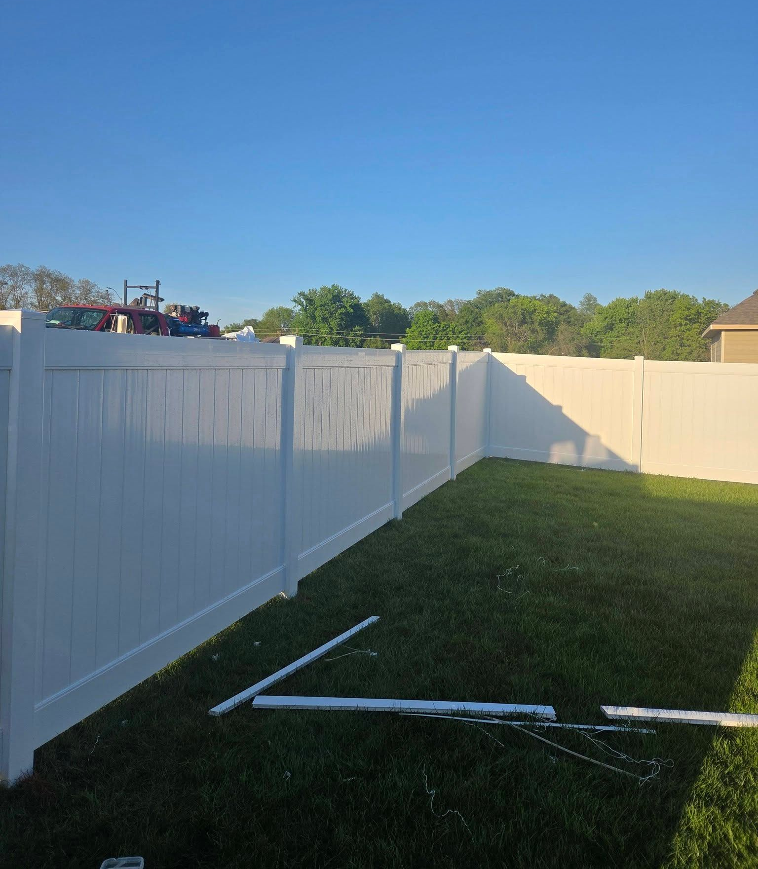 White vinyl fence bordering a grassy backyard under a clear blue sky, with fence panels on the ground.