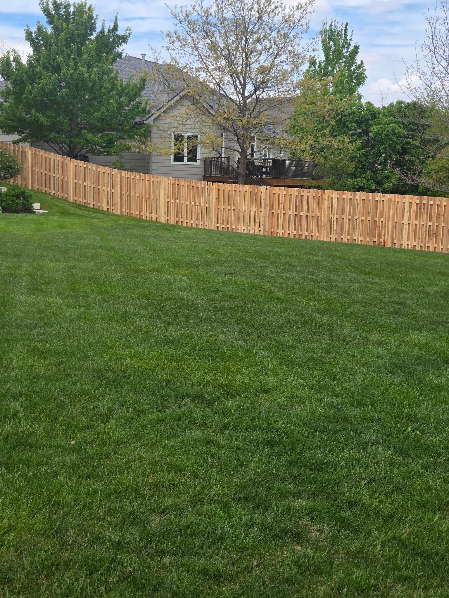 Backyard with green lawn, wooden fence, and trees in front of a house under a partly cloudy sky