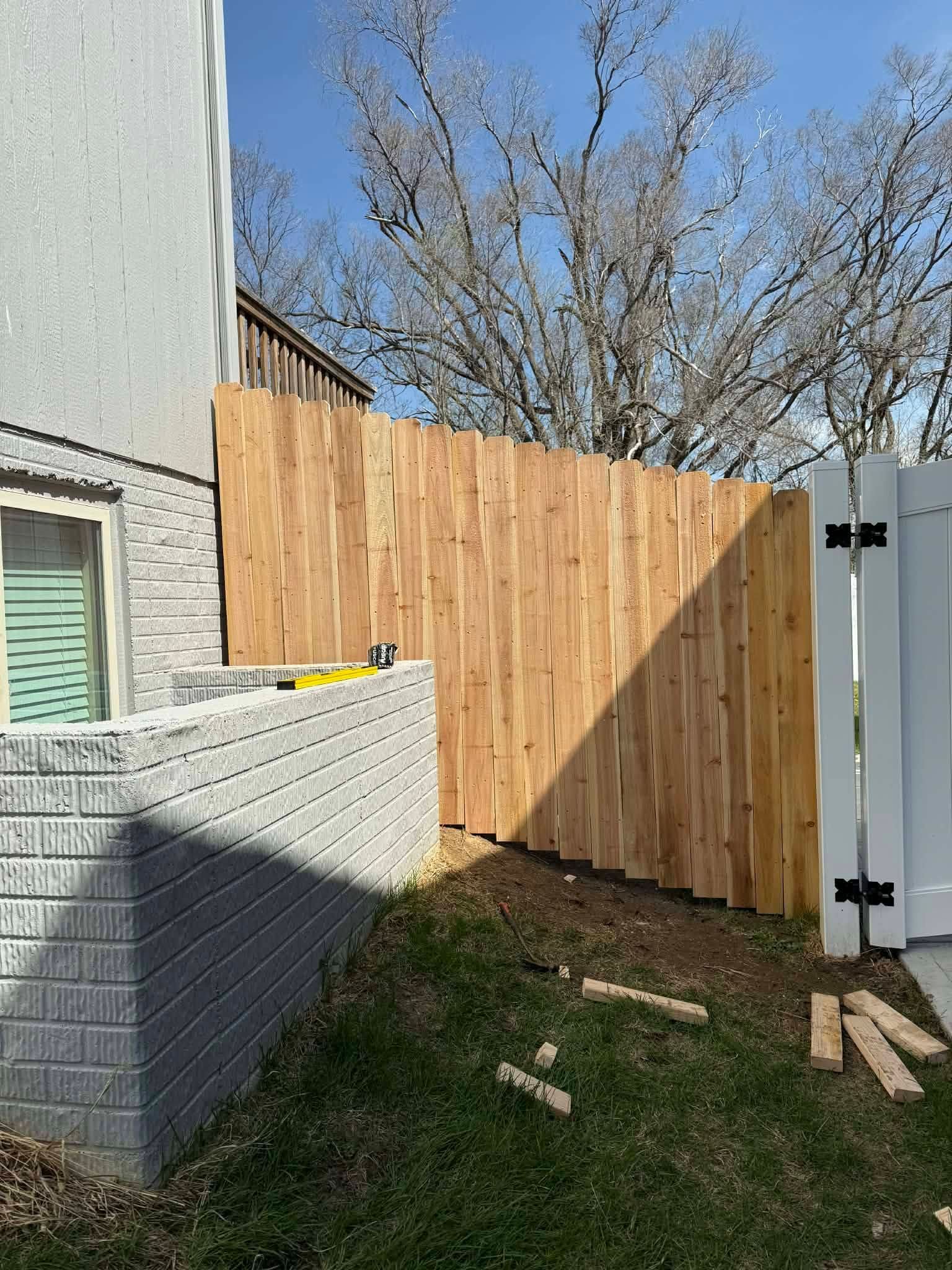 Backyard corner with wooden fence, gray house wall, white gate, and patchy grass in sunlight