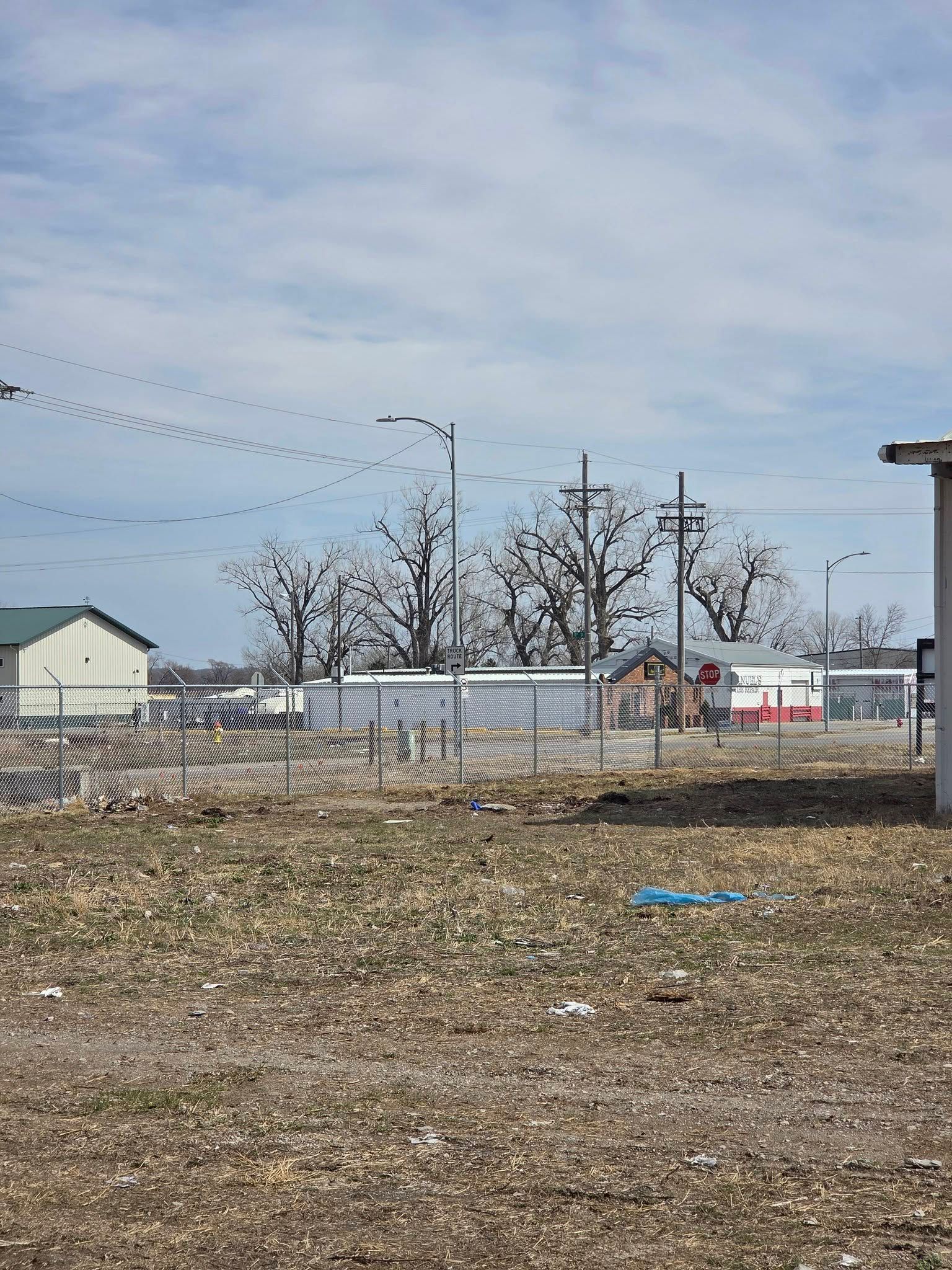 Empty gravel lot with chain-link fence, utility poles, and buildings under a cloudy sky.