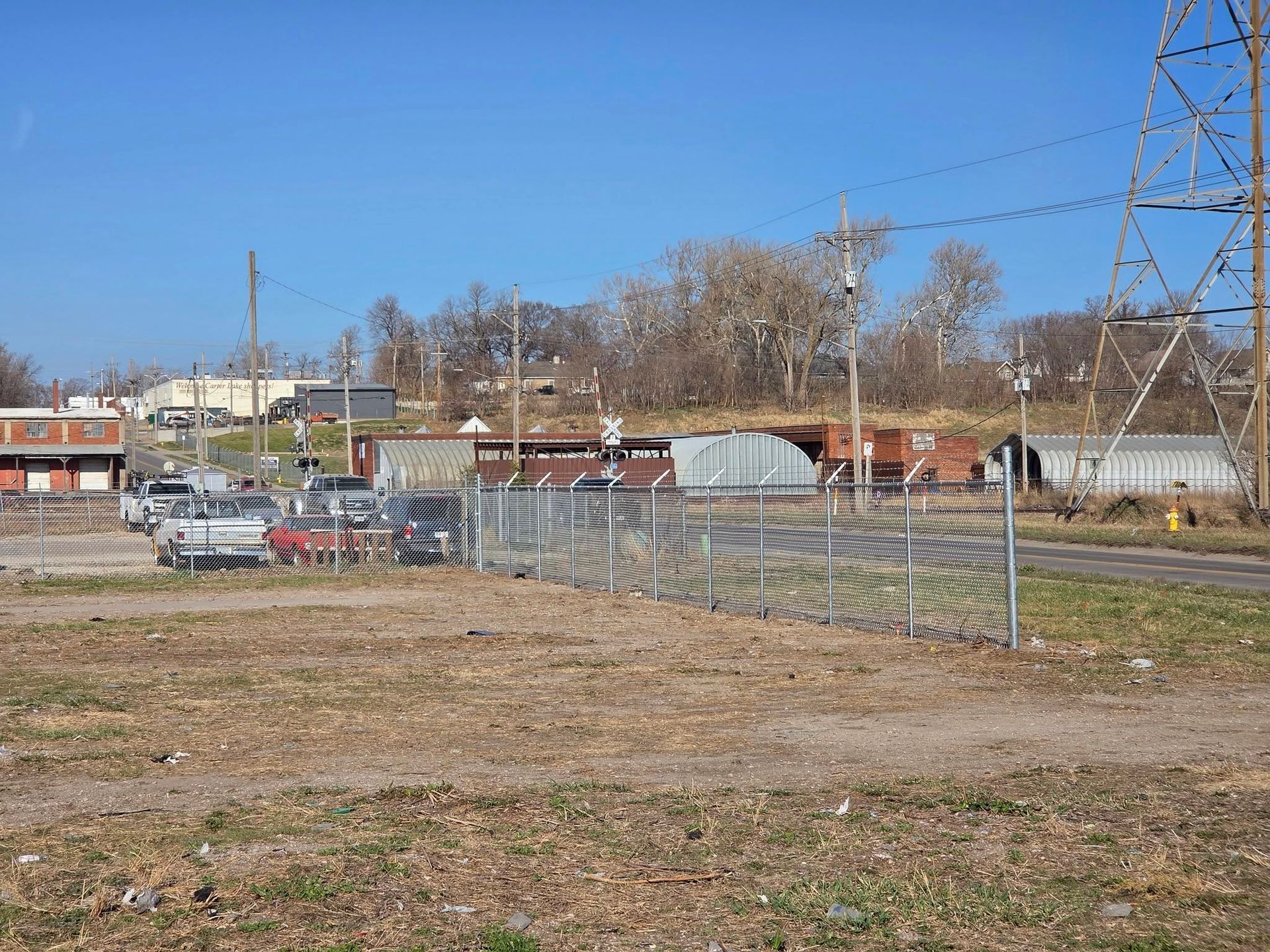 Rural rail yard with chain-link fence, parked cars, freight cars, and power lines under a clear blue sky