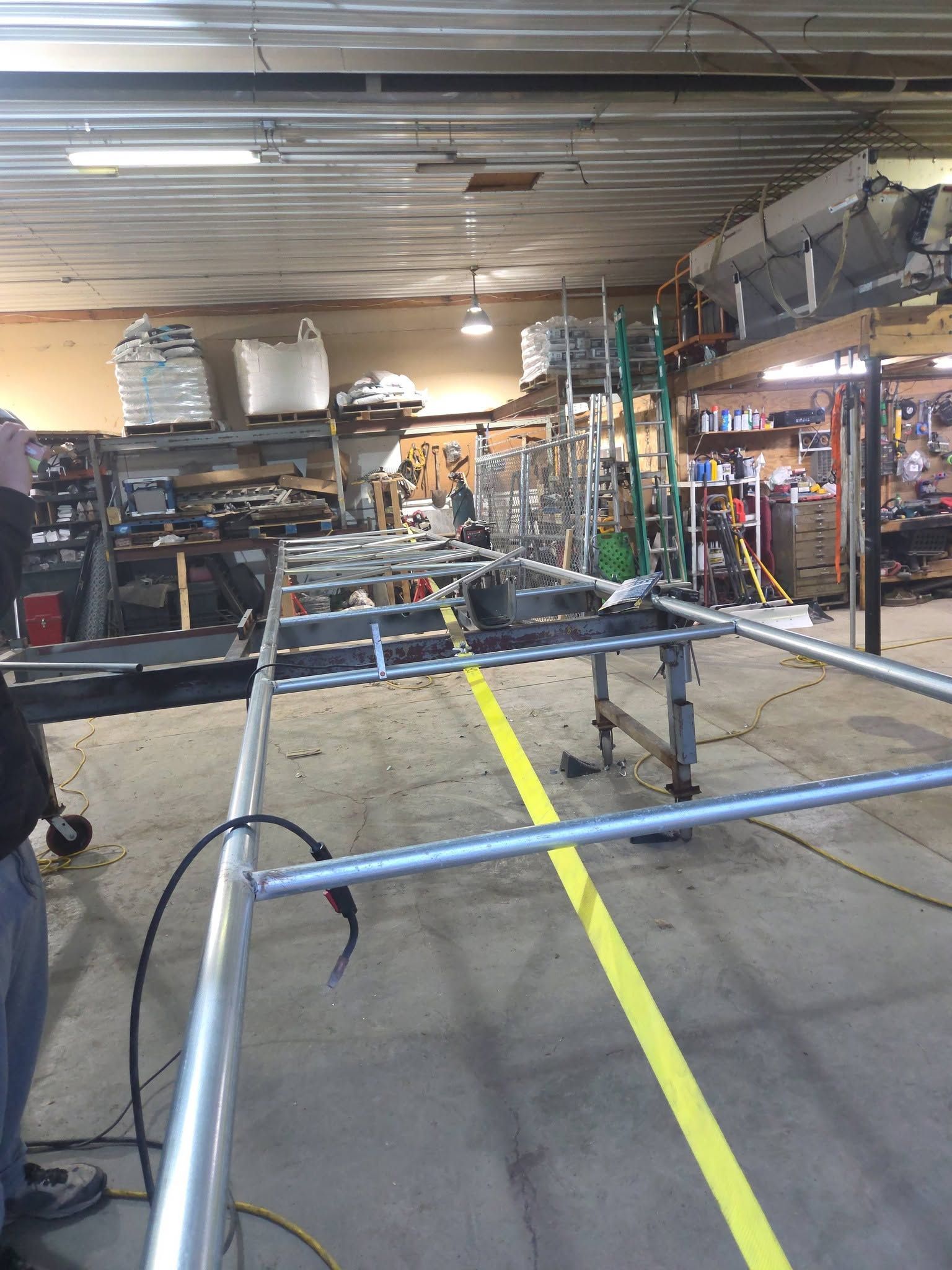 Indoor auto shop with stacked car frames, tools, and a yellow floor line under bright fluorescent lights