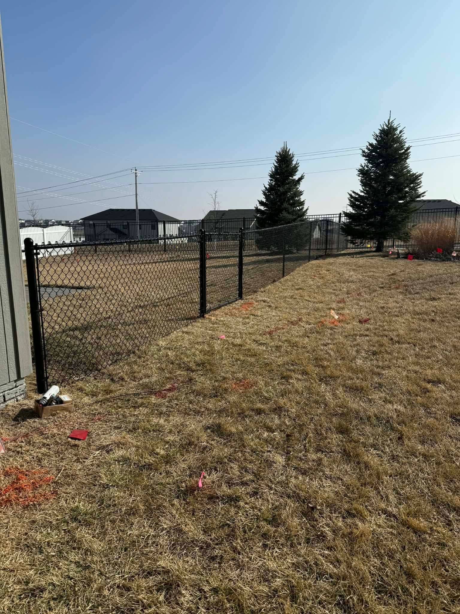 Chain-link fence beside a dry grassy field with trees and a house under a clear blue sky.