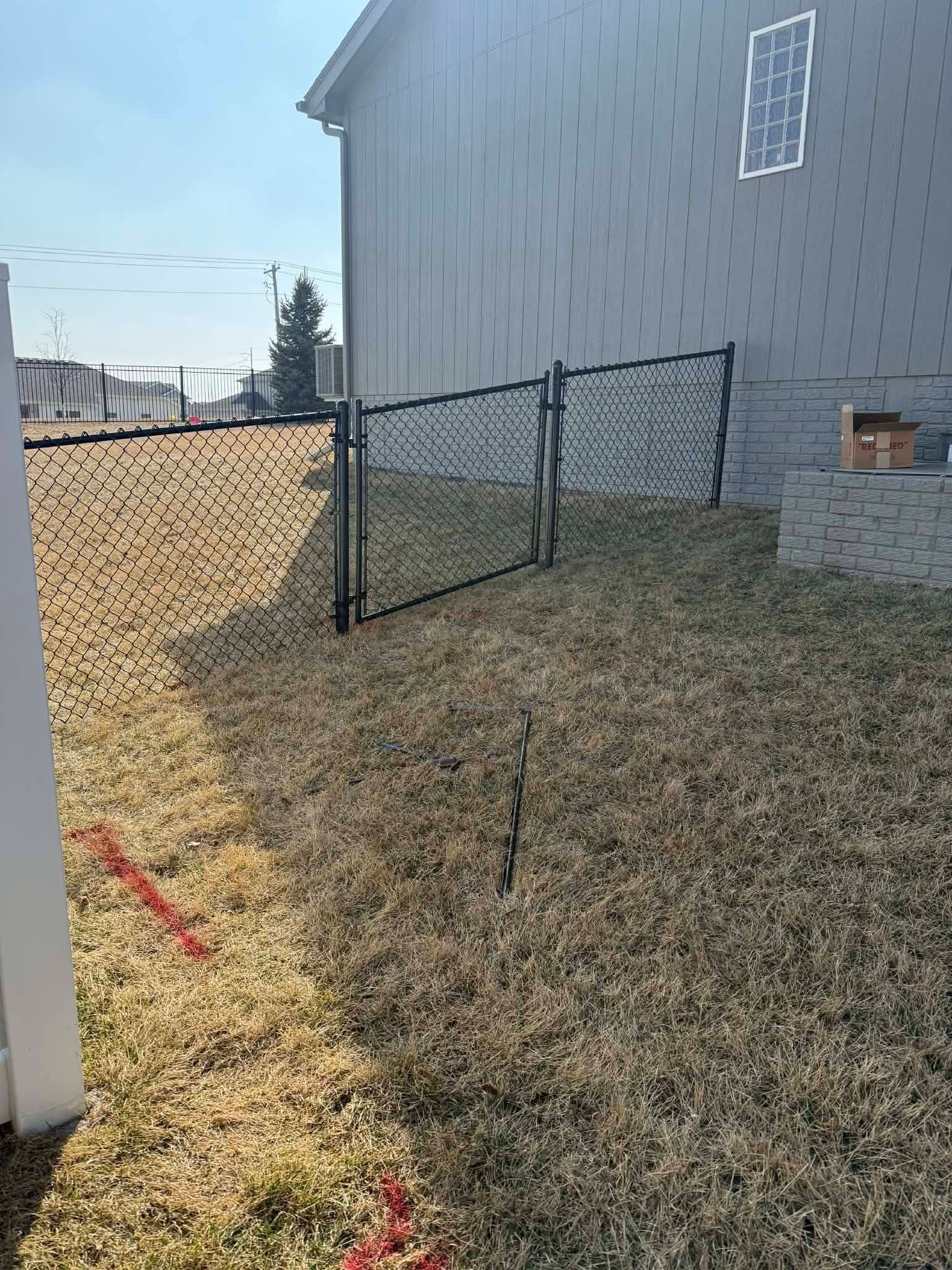Chain-link fence in a dry backyard beside a gray house under bright sunlight.