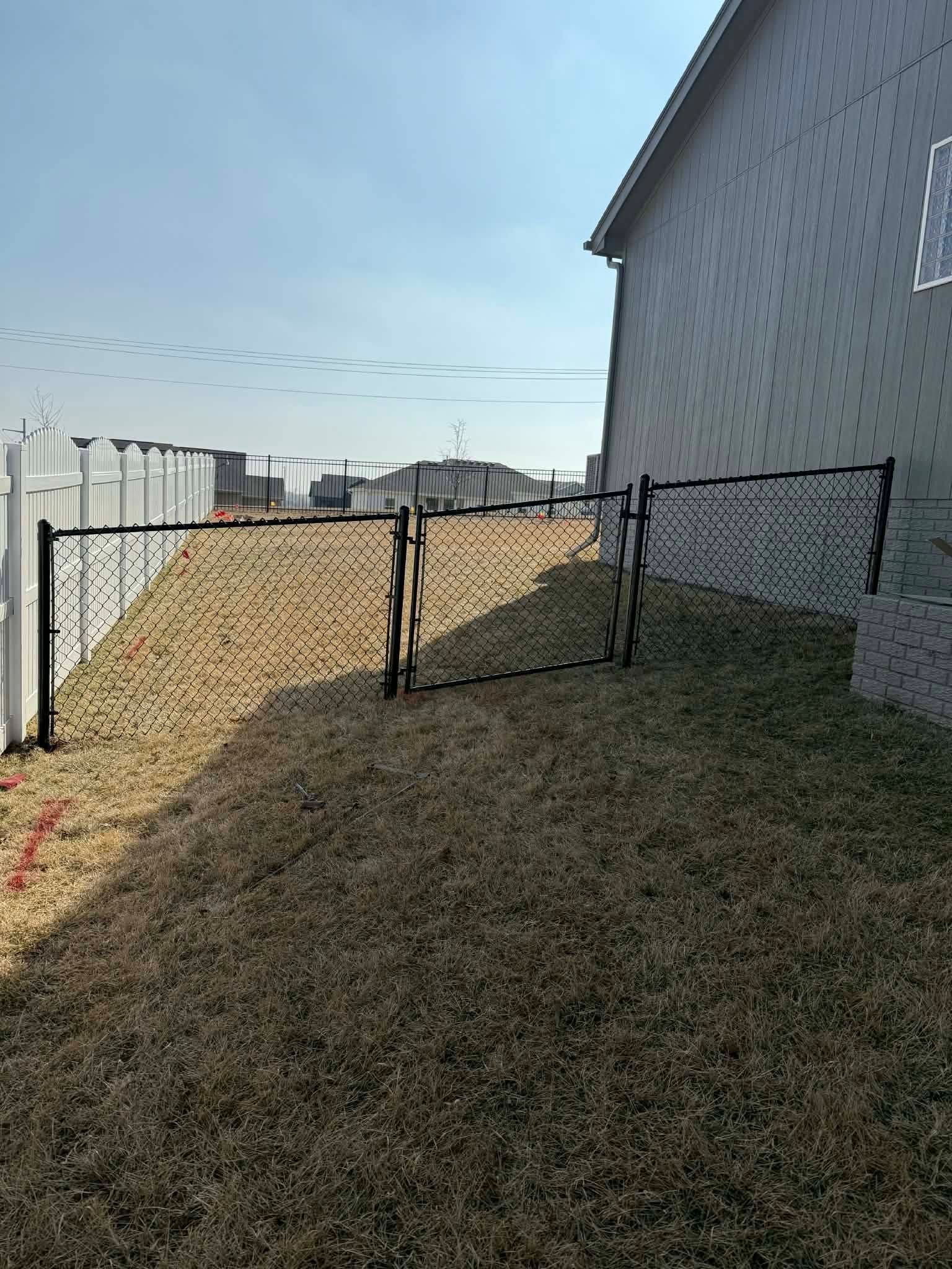 Narrow grassy side yard beside a house, with chain-link fences and a white fence along the left side.