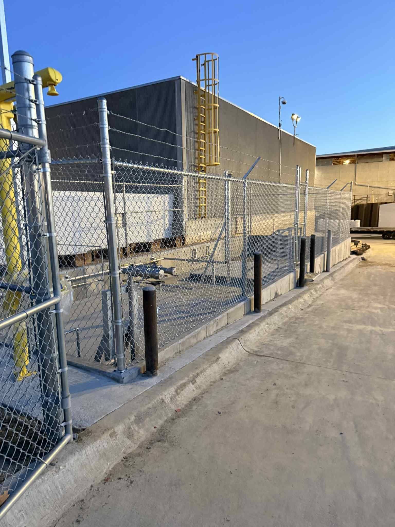 Chain-link fenced industrial alley with utility poles and a concrete building under a clear blue sky