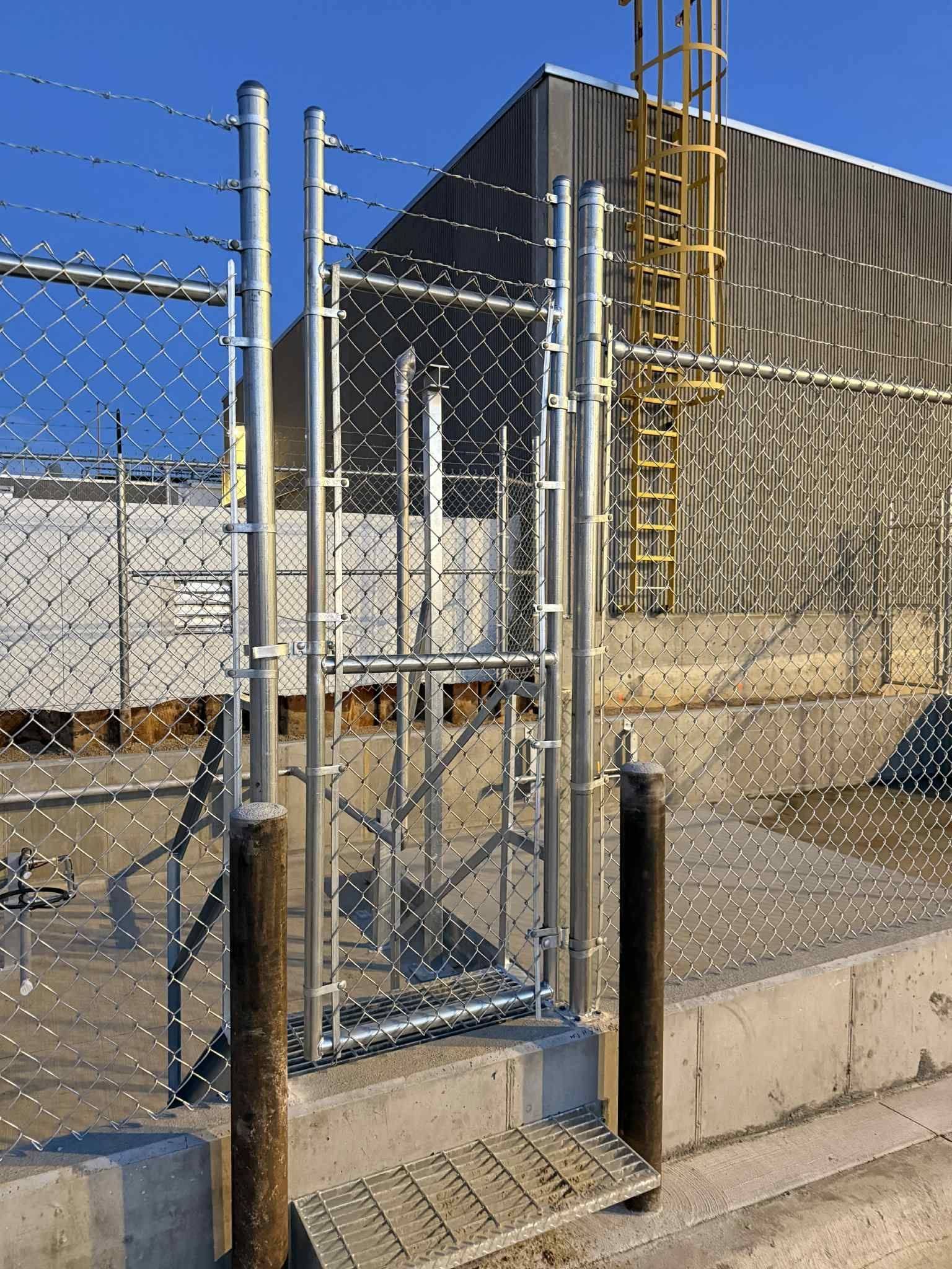Chain-link gate with barbed wire in front of an industrial building under a blue sky