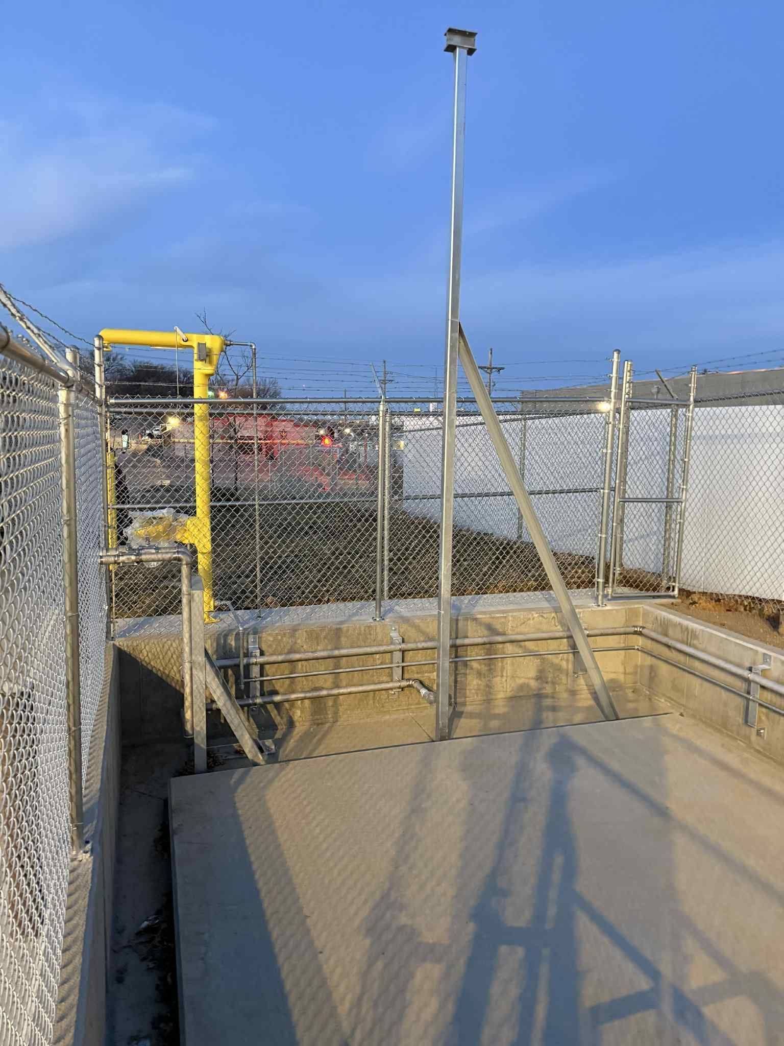Chain-link fenced industrial area with a yellow ladder and tall pole over a concrete pit.