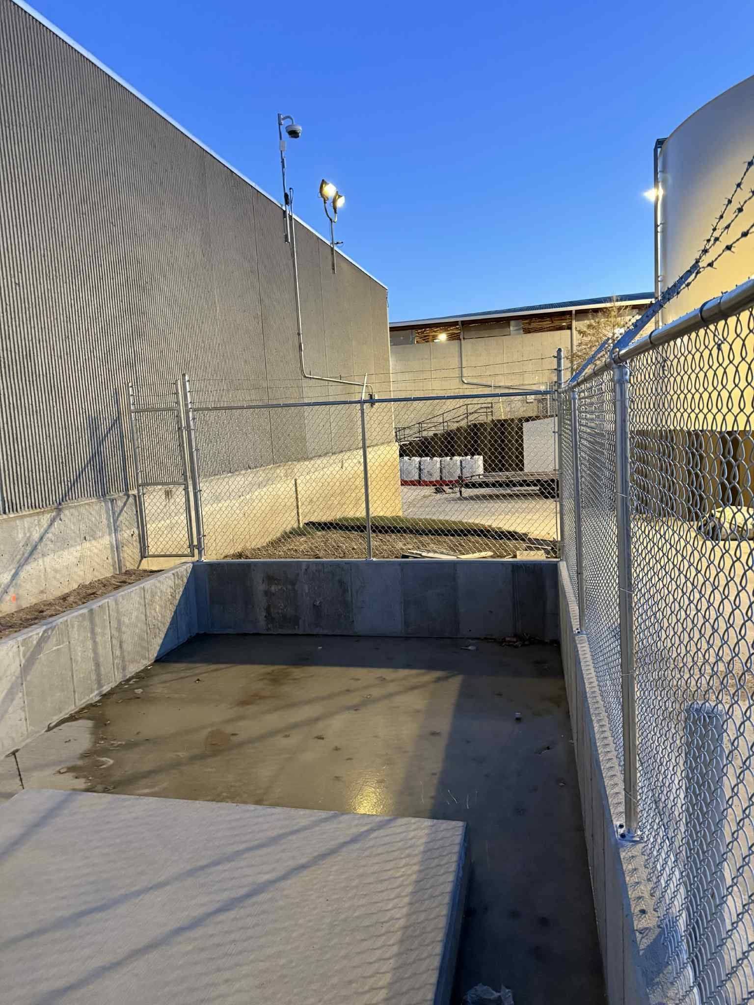Empty concrete yard between chain-link fences in bright sunlight, with a rooftop view and a concrete bench in front