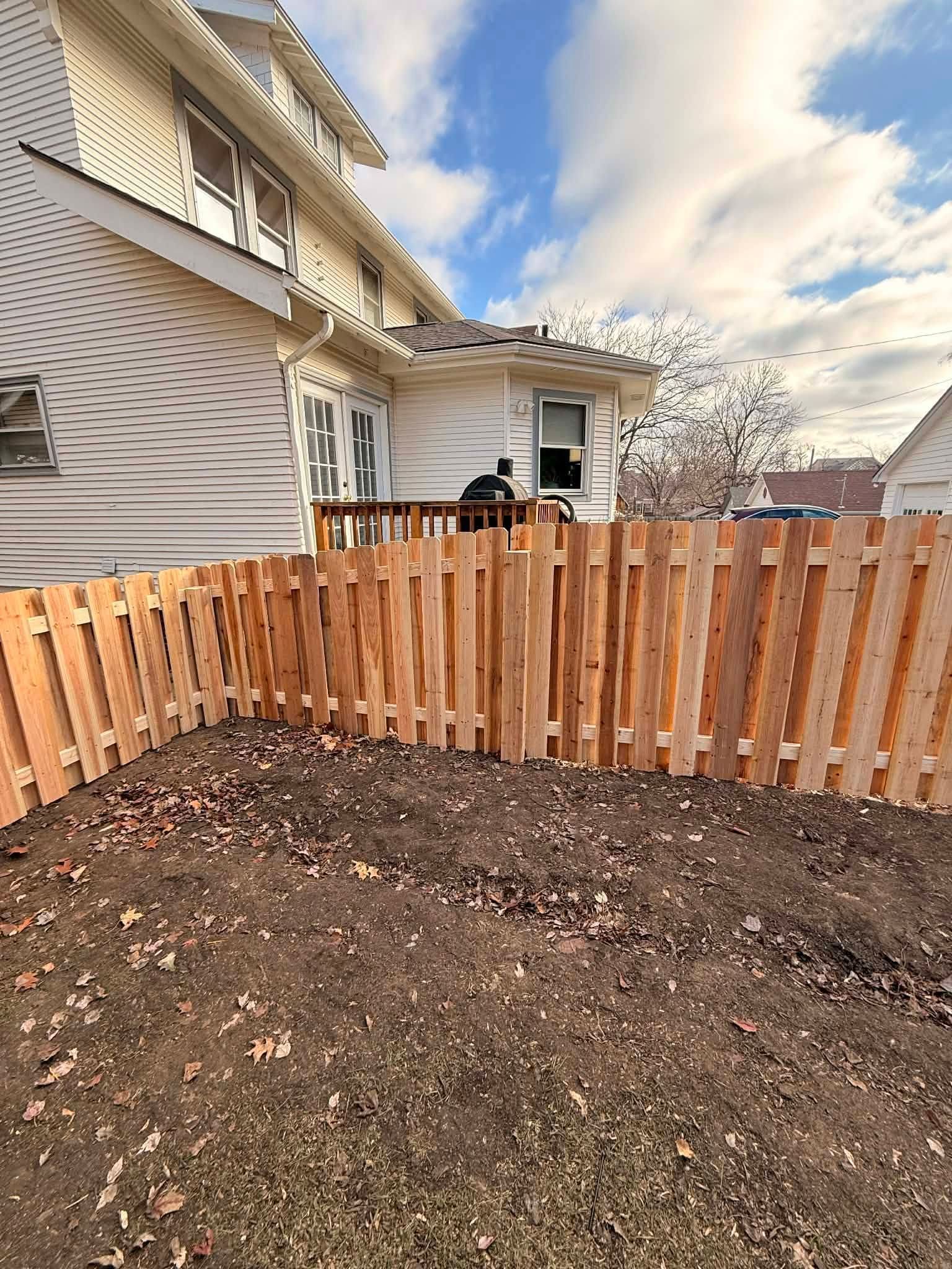 Backyard with a wooden fence, bare soil, and a house with a covered patio under a cloudy sky.