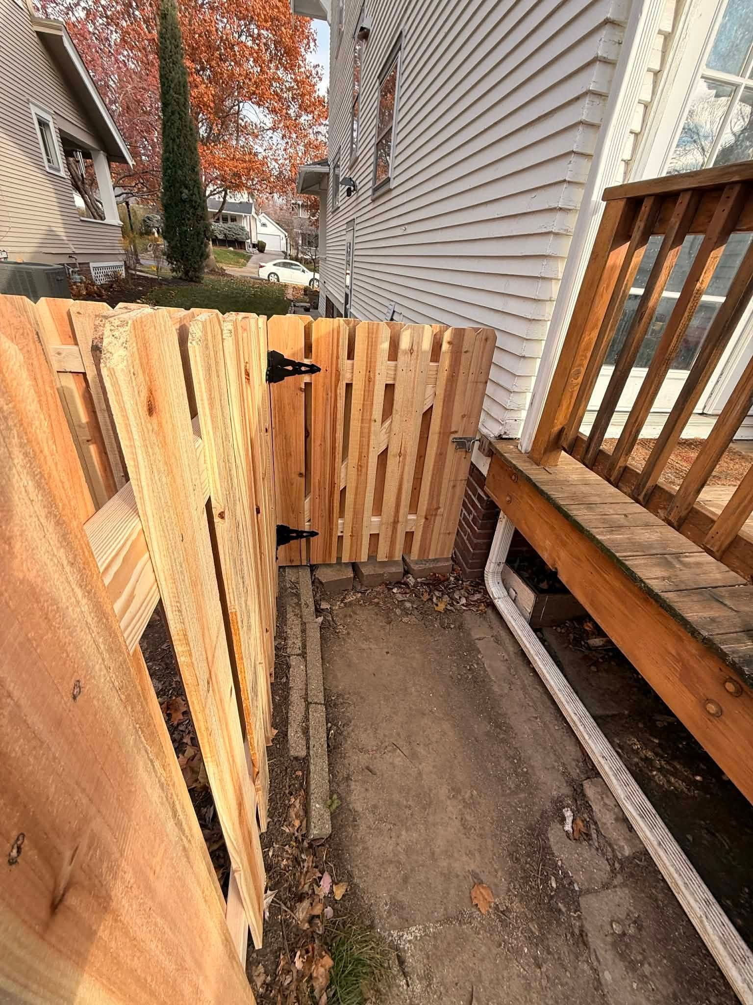Narrow fenced walkway beside a house, with unfinished wooden gates and autumn trees beyond.