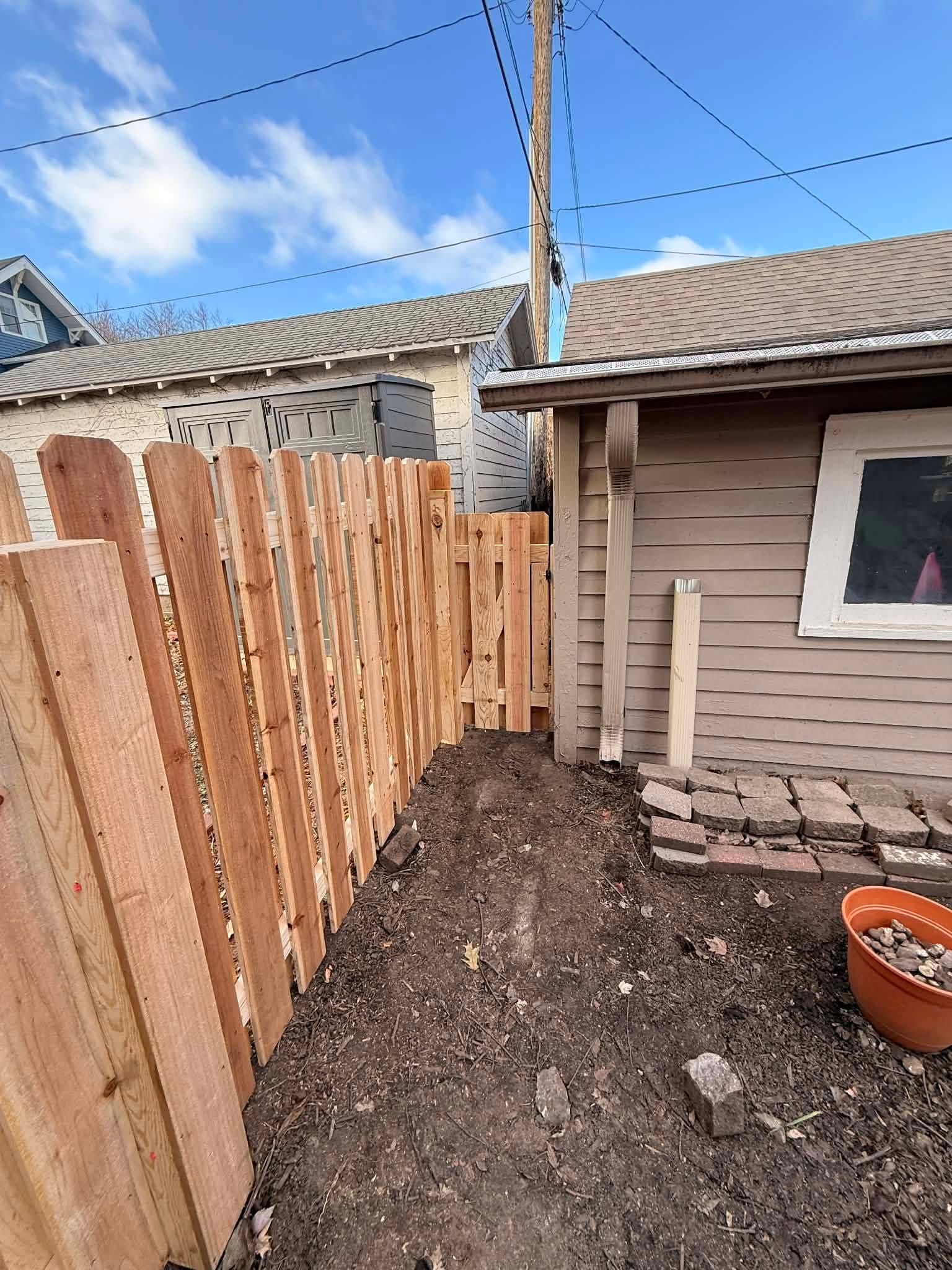 Narrow backyard path with a wooden fence, dirt ground, and beige house under a blue sky.