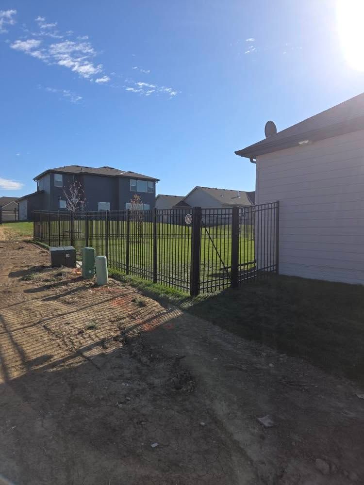 Residential street with fenced yard, gravel driveway, and blue two-story house under a bright sky