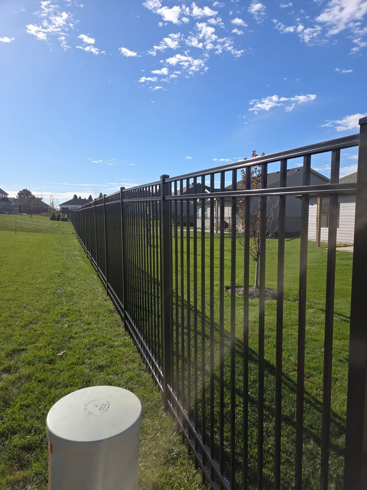 Black metal fence beside a green field under a bright blue sky with scattered clouds.