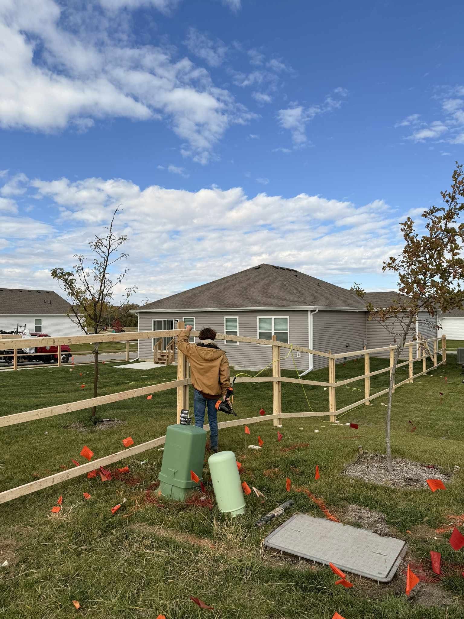 Person watering a young tree in a grassy yard beside a house on a cloudy day