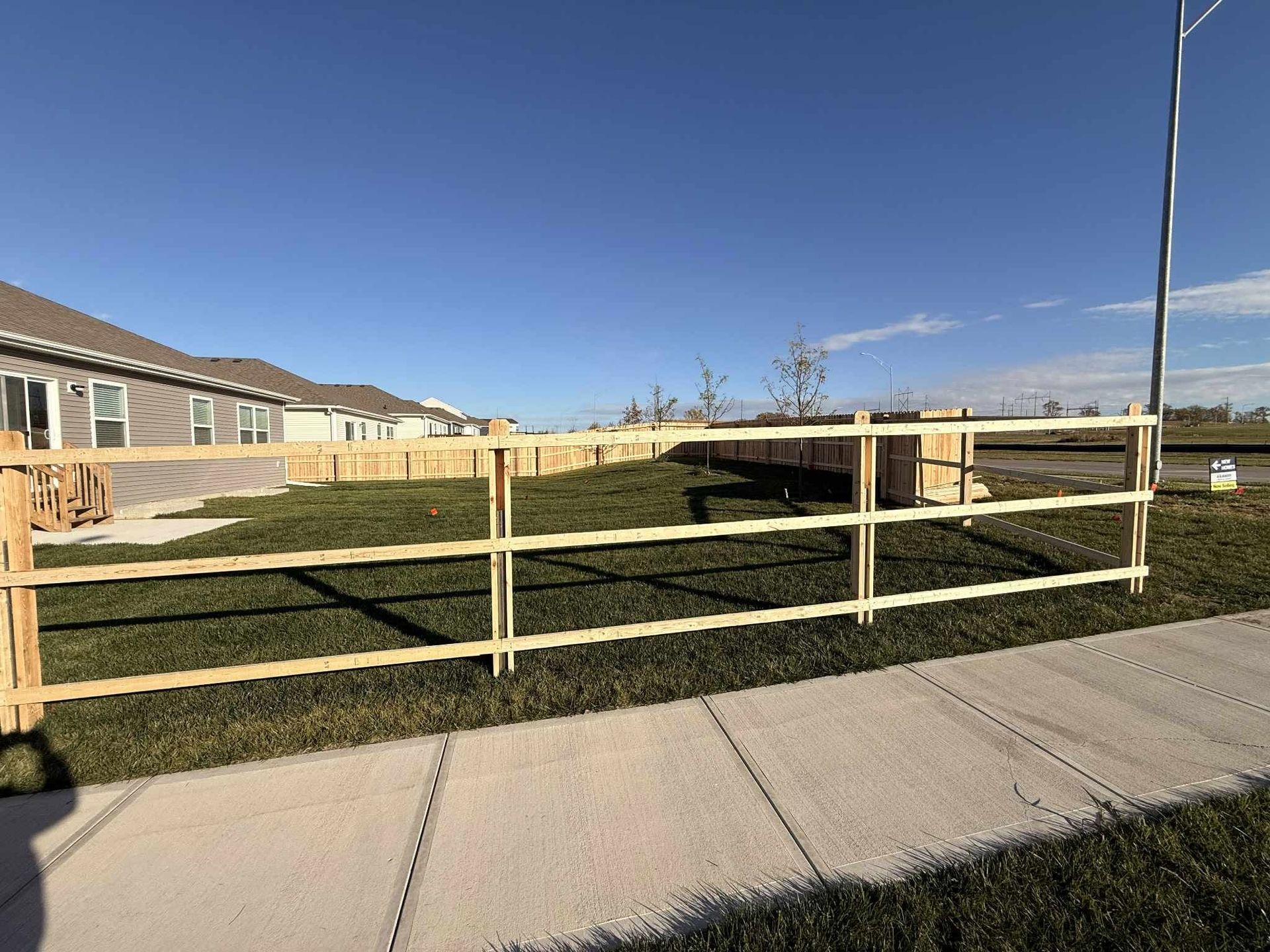 Wooden fence along a grassy roadside beside a sidewalk and brick homes under a clear blue sky