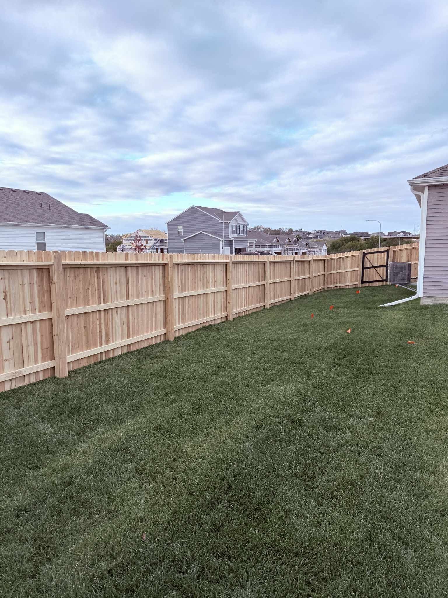 Backyard with a wooden fence, green lawn, and cloudy sky, with houses in the distance