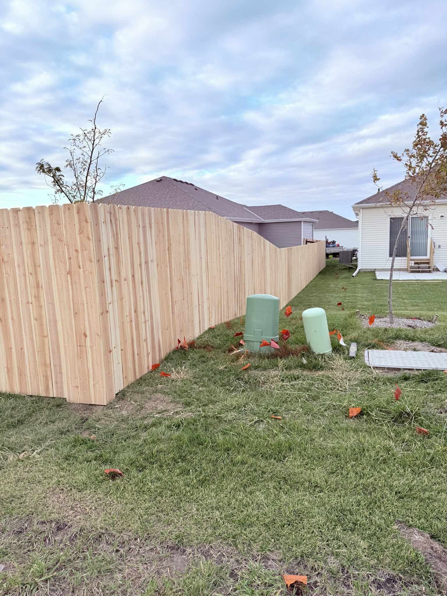 Backyard with a wooden fence, green utility boxes, and a house in the background under a cloudy sky