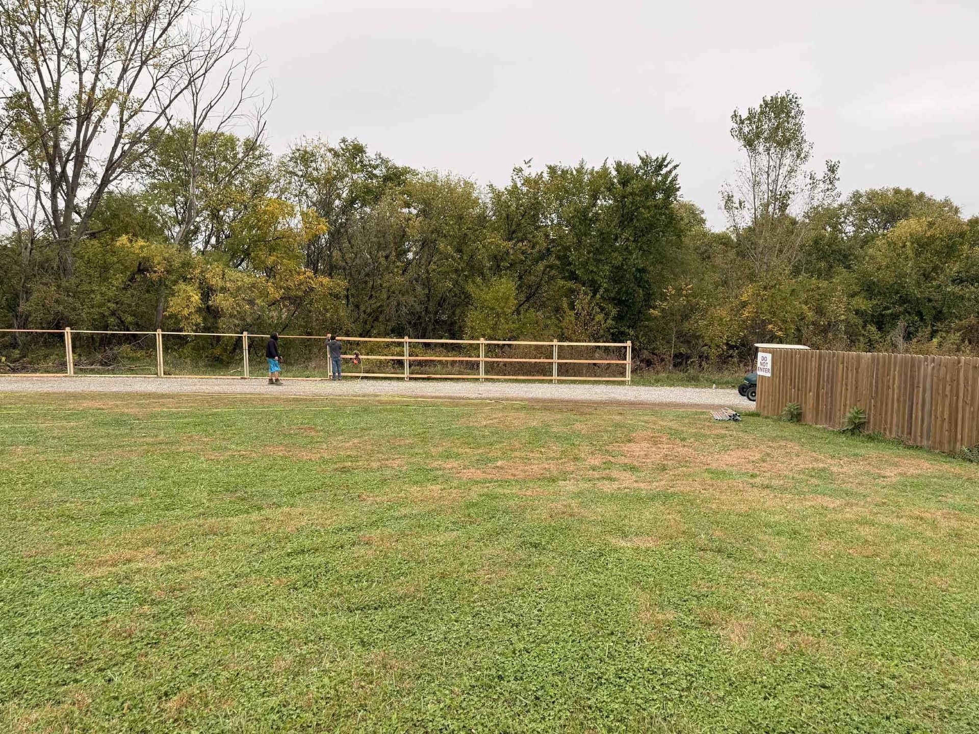 Grassy fenced park with a small trash bin and trees in the background under an overcast sky