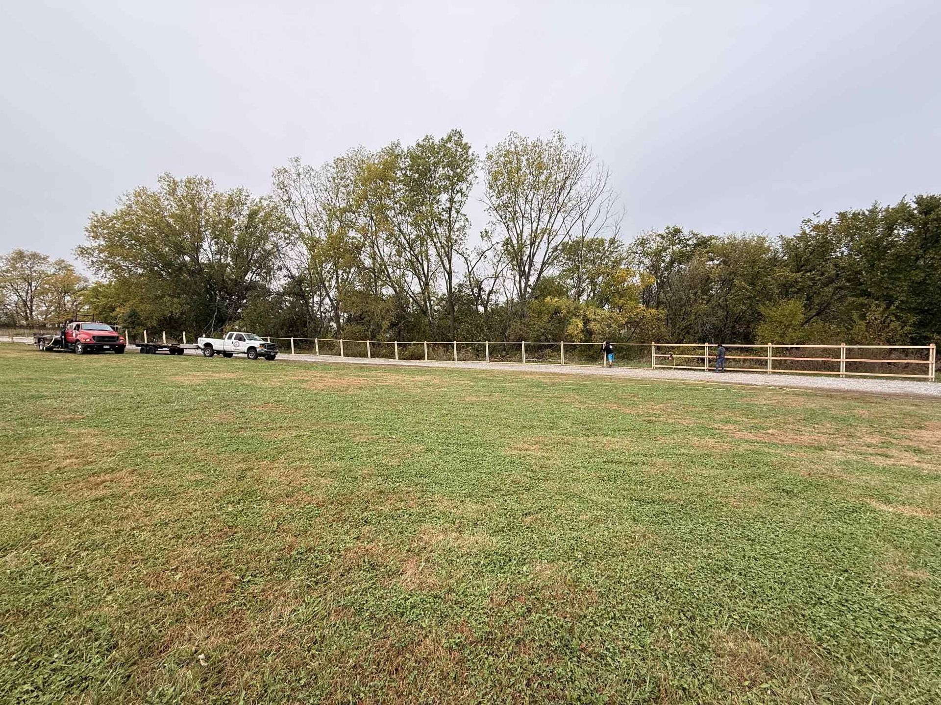 Grassy park field beside a white fence and trees, with parked cars under a cloudy sky