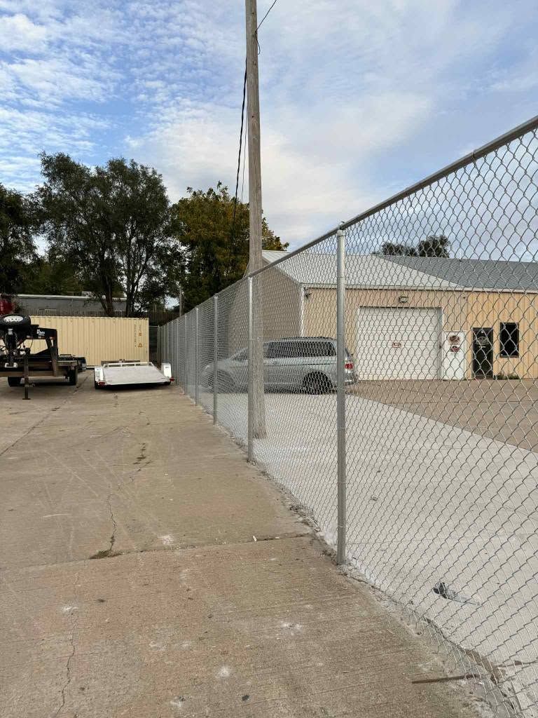Chain-link fence beside a paved lot with a white building and parked utility cart under a cloudy sky