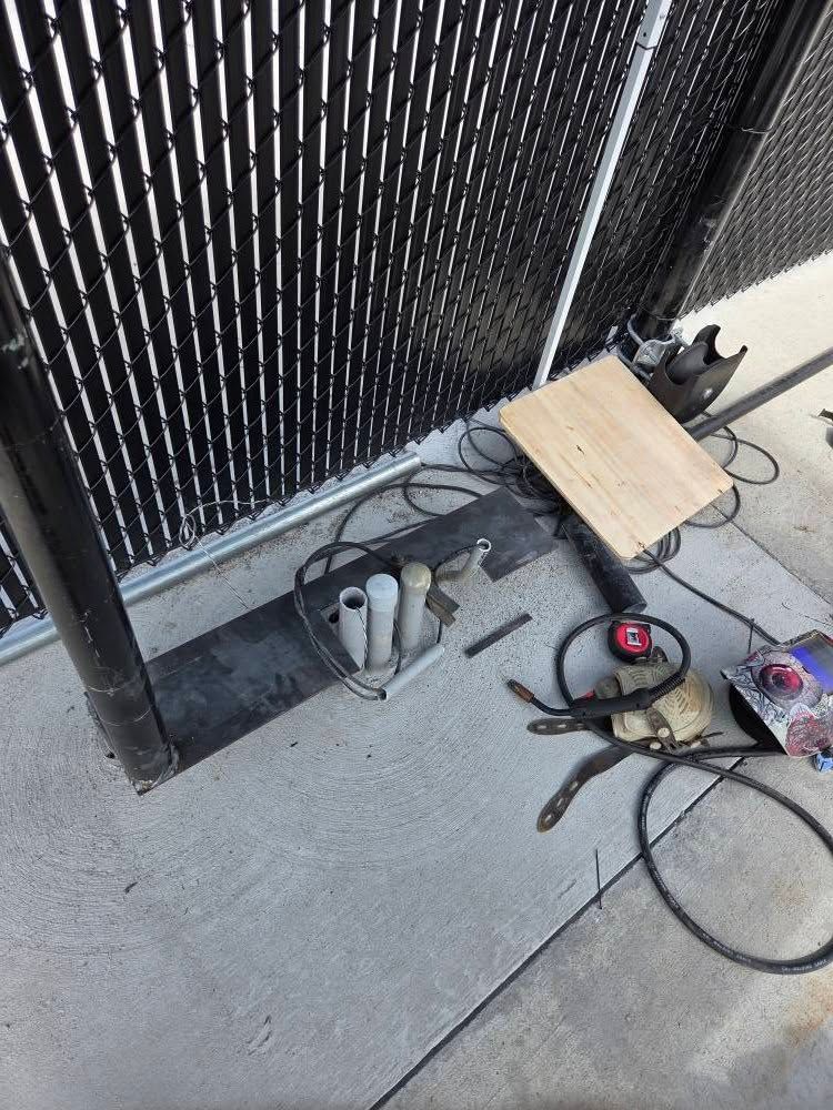 Broken pavement by a black fence with a wooden board, cables, and tools scattered on the ground