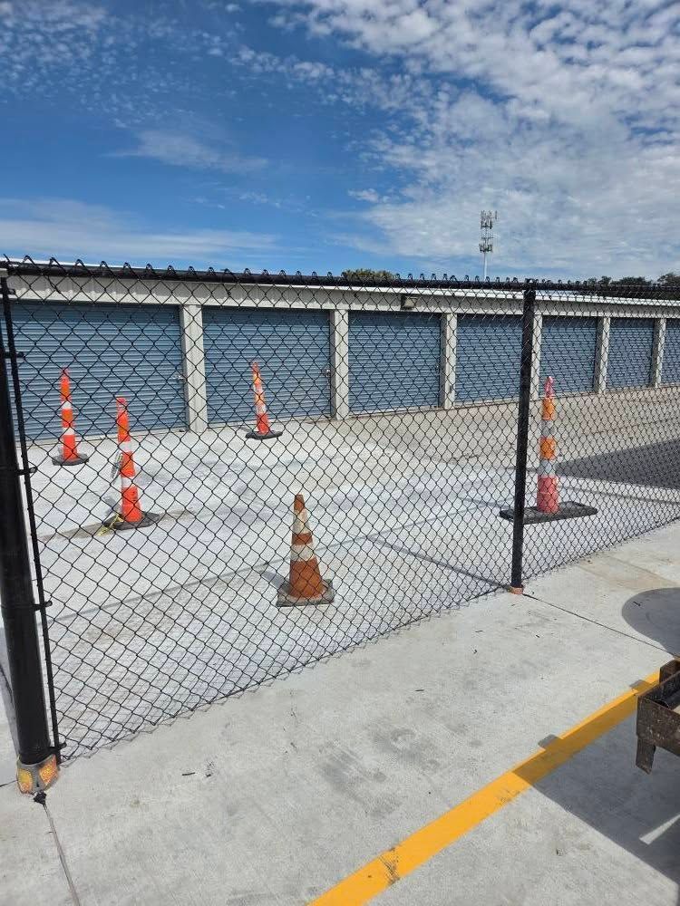Orange traffic cones behind a chain-link fence outside storage units under a blue sky