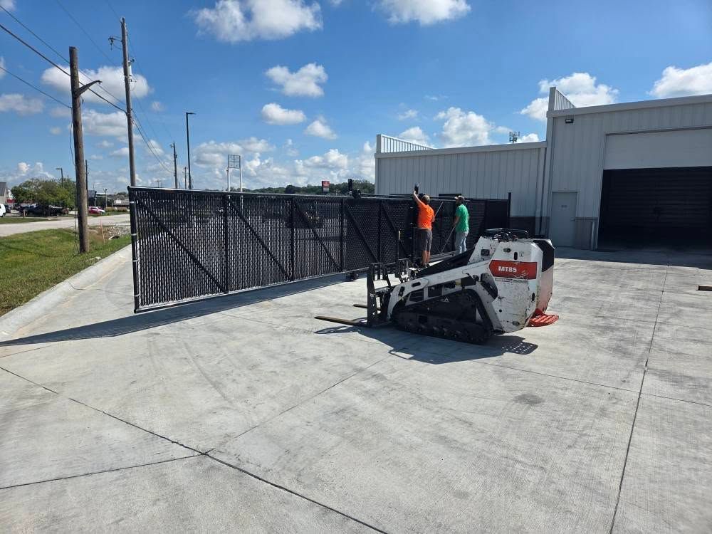 Workers beside a white skid steer near a black fence outside a warehouse under a blue sky