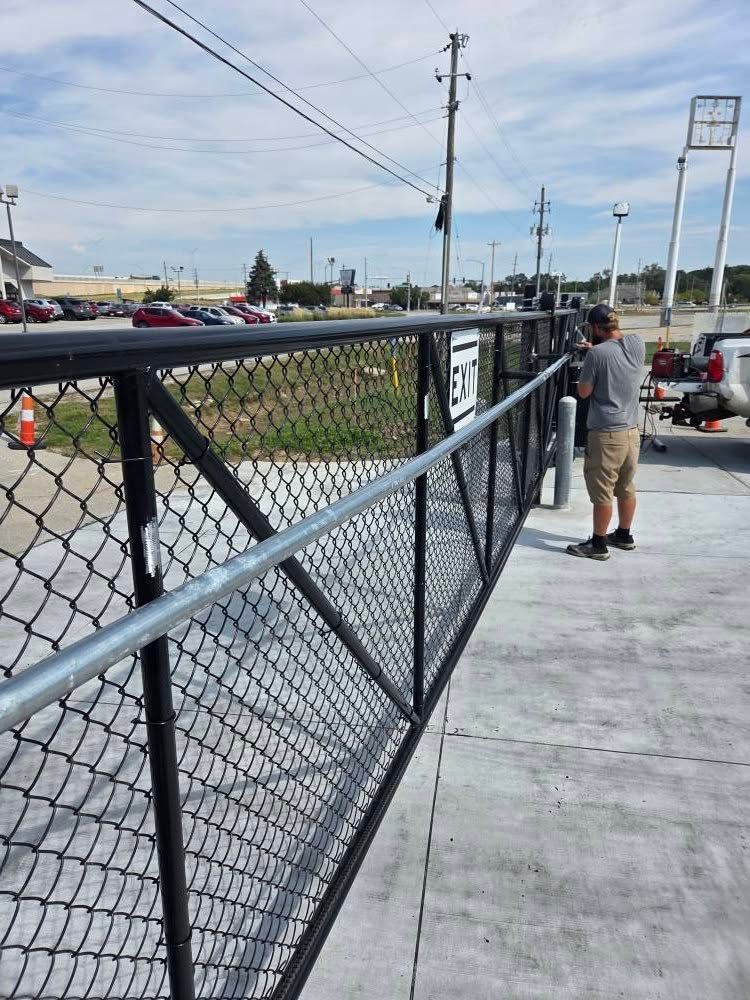 Person leaning on a chain-link fence beside a paved walkway, overlooking a road and parking lot.