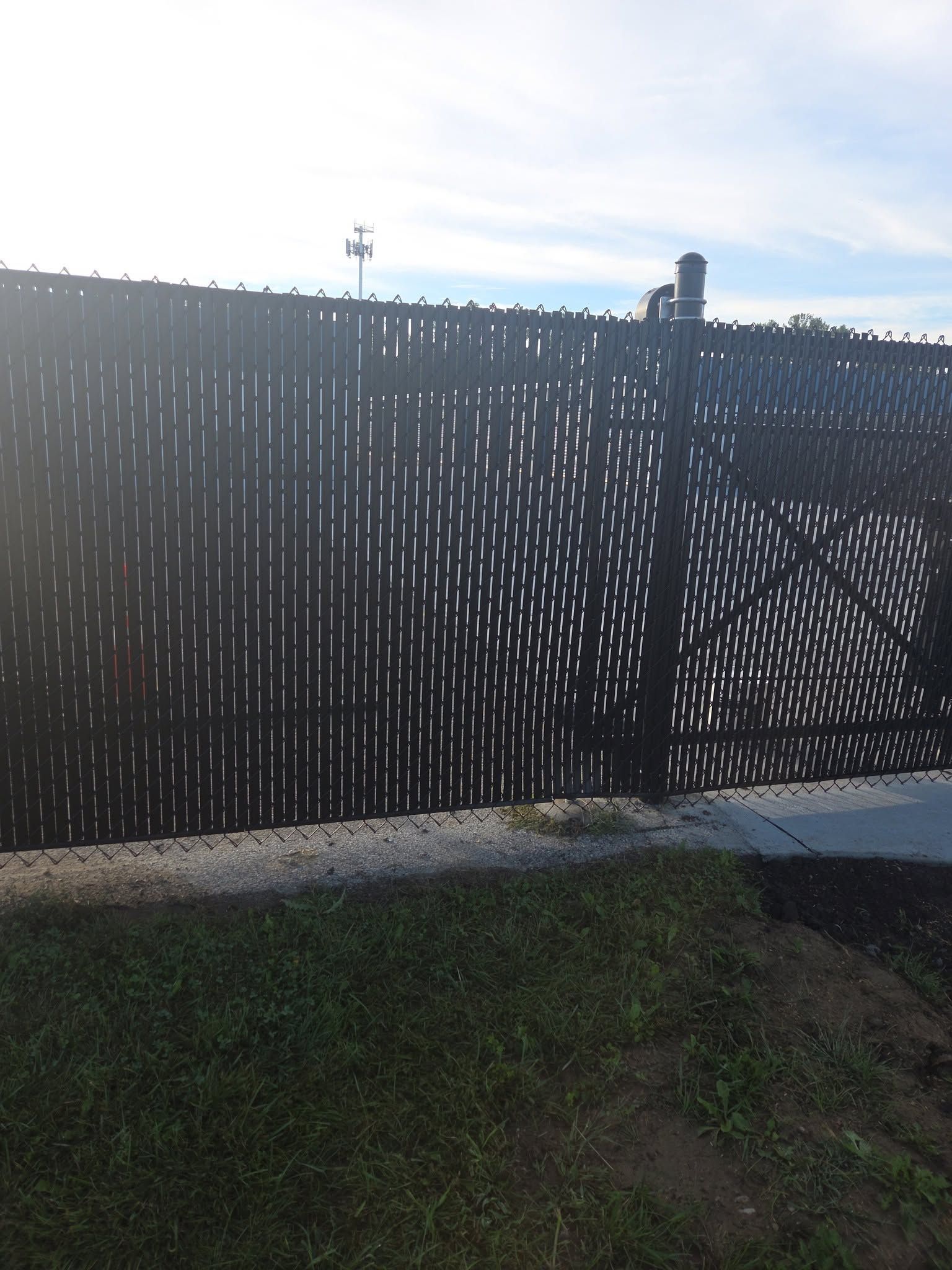 Black privacy fence with a gate beside a concrete path and grass under a bright sky