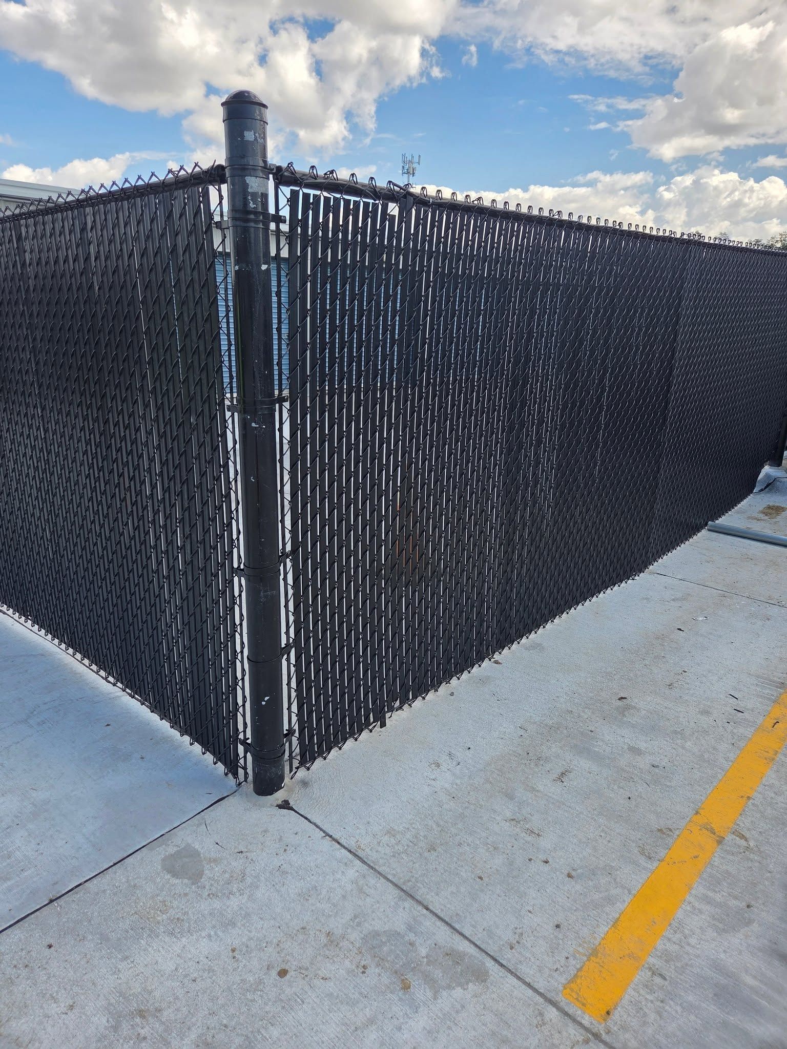 Black chain-link fence corner beside a concrete sidewalk with a yellow curb line under a cloudy sky