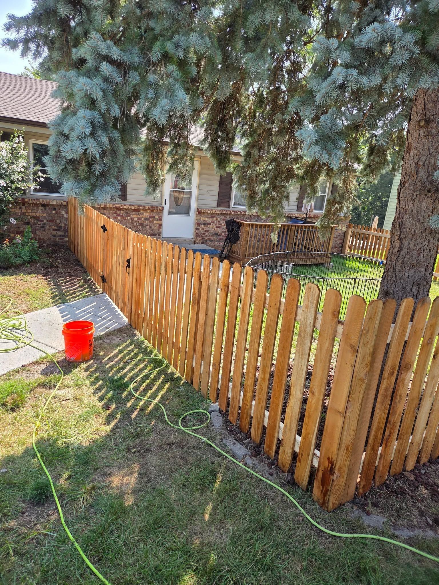 Wooden picket fence around a yard with trees, a house, and a red bucket on the grass