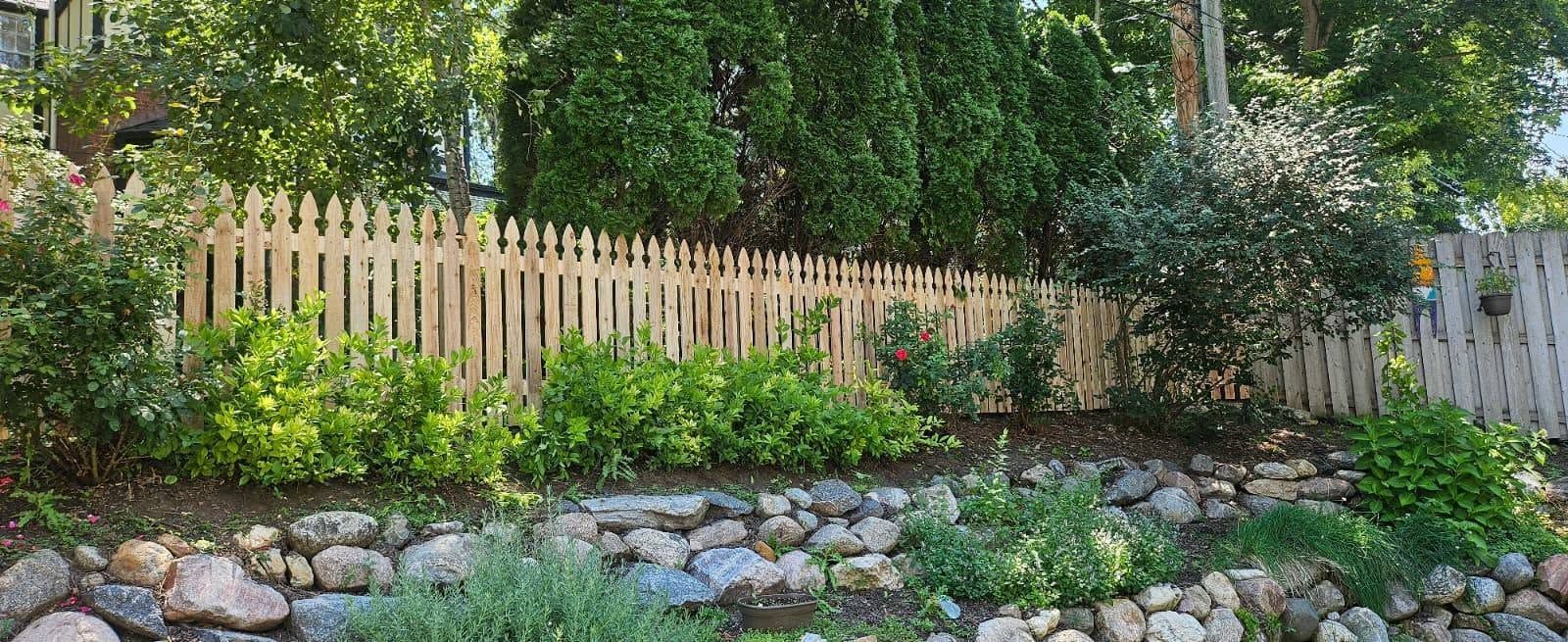 Wooden fence behind a lush garden bed with rocks and green shrubs