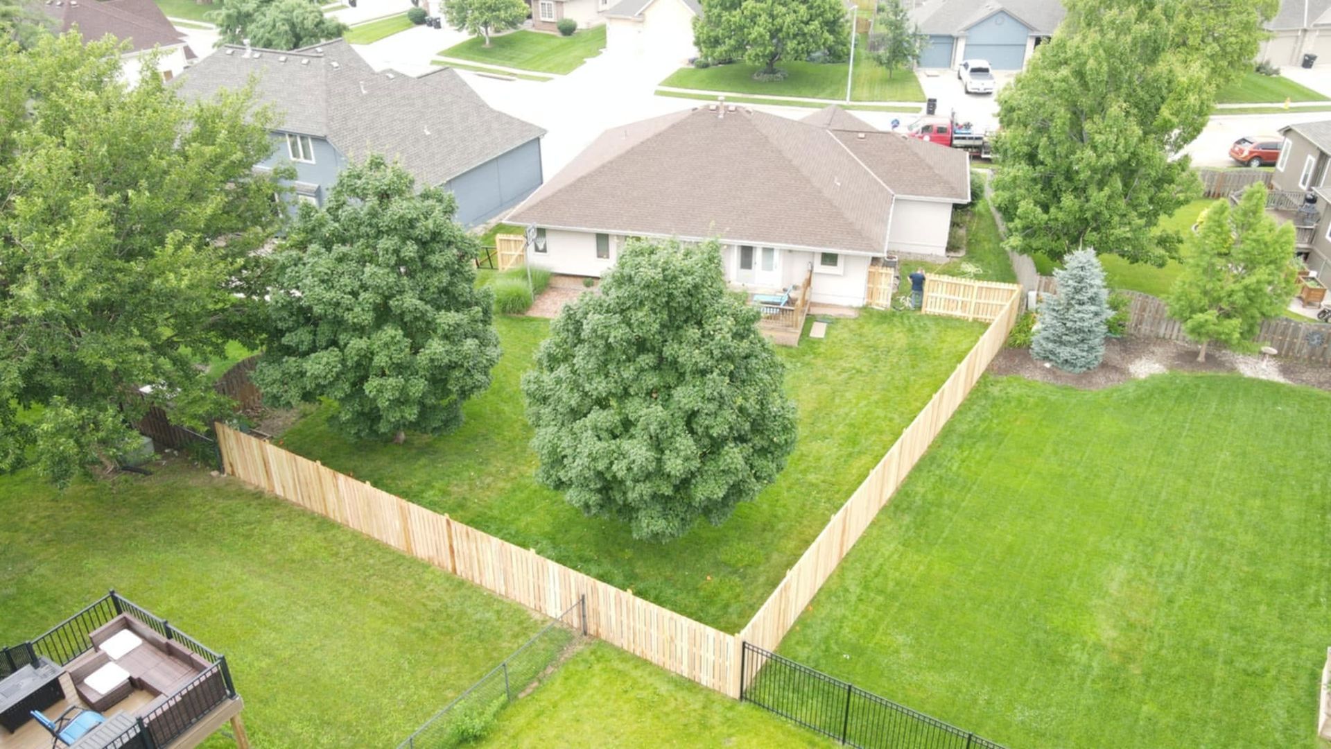 Aerial view of a fenced backyard with a large tree, lawn, and suburban houses nearby.