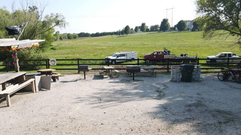 Outdoor picnic area with benches, a gravel foreground, and a grassy field under bright sunlight.