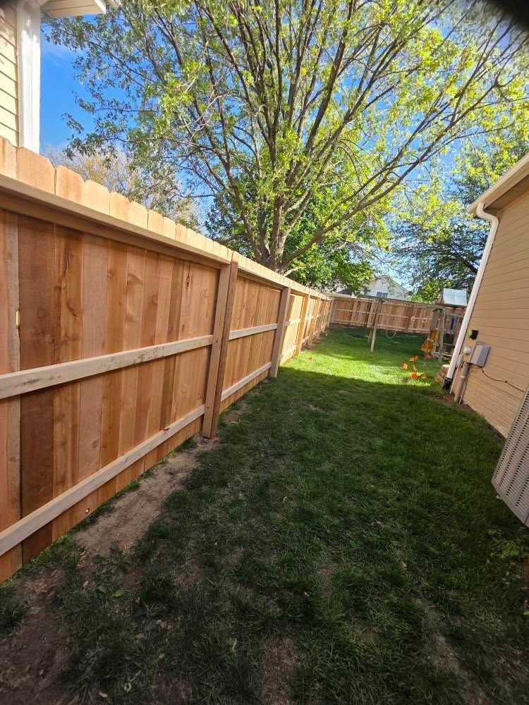 Narrow grassy backyard between a wooden fence and house, with a large tree at the end.
