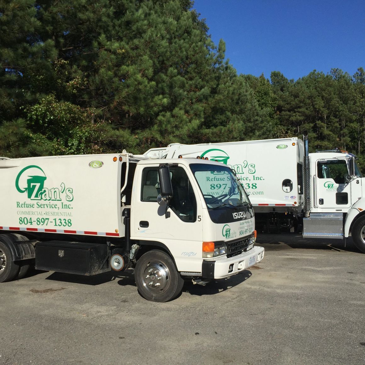 Two white Zan's Refuse Service, Inc. garbage trucks parked in a gravel lot against a backdrop of trees.