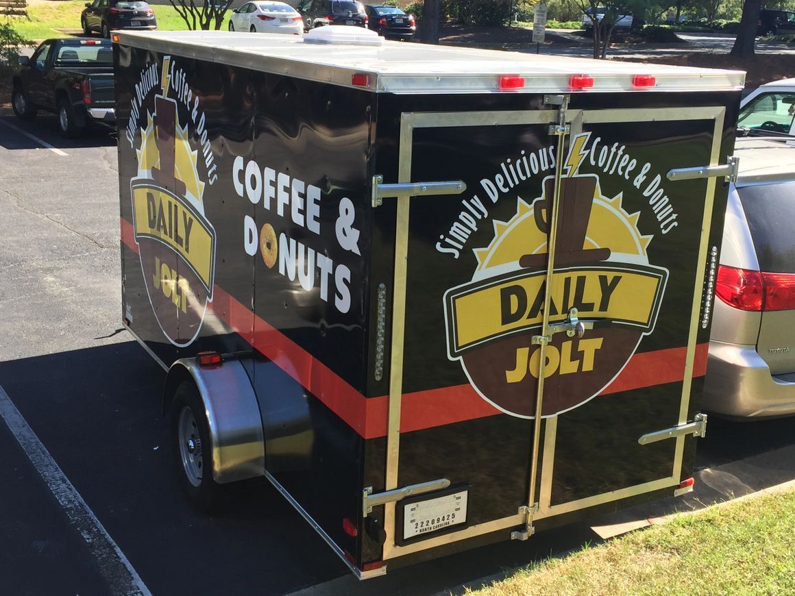 A black Daily Jolt trailer with red stripes, parked outdoors, advertising coffee and donuts on its side and back doors.