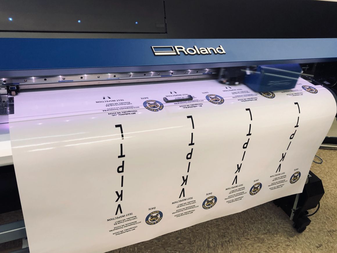 A large-format Roland printer printing vertical text and logos onto a continuous roll of white vinyl material.