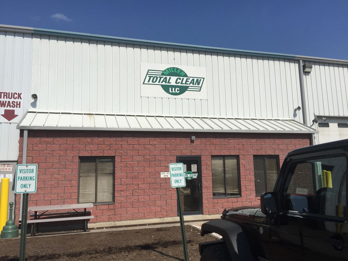 A red brick and white metal building with a Total Clean LLC sign, a picnic table, and a partial view of a black vehicle.