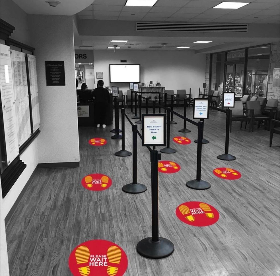A waiting area with red social distancing floor decals and black stanchion posts guiding a queue toward a service counter.