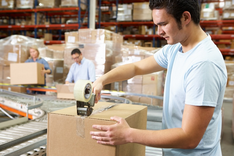 A man is taping a cardboard box in a warehouse.