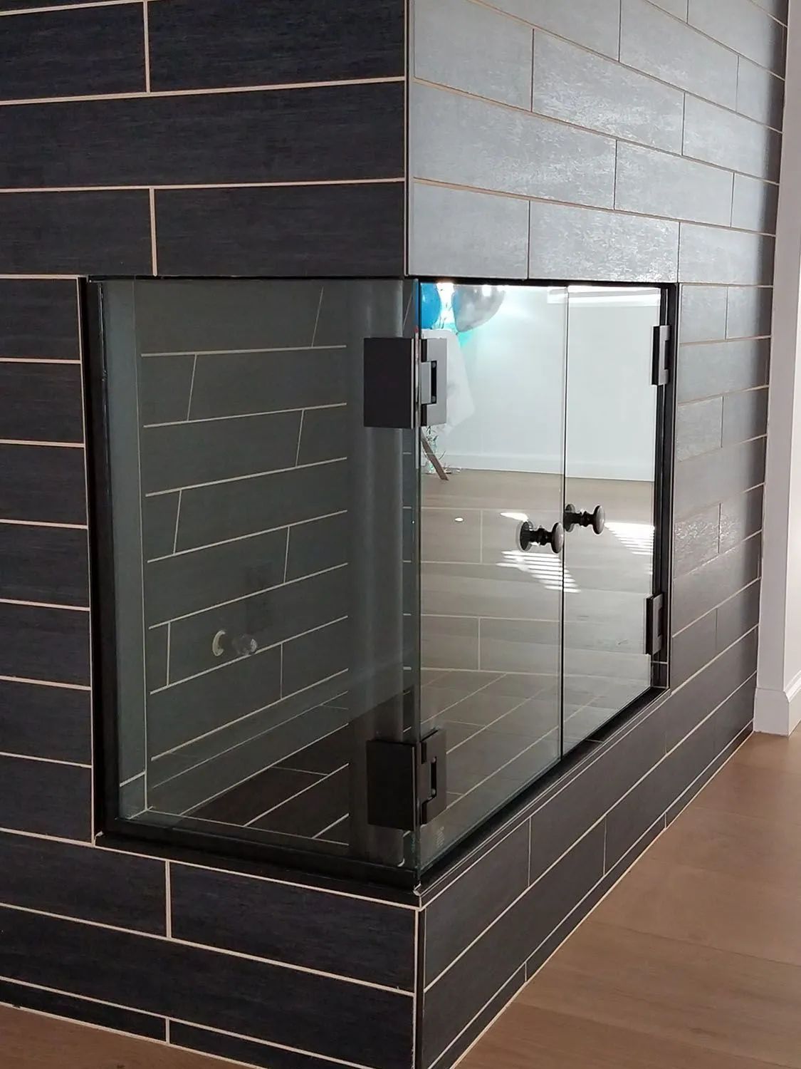 Black corner fireplace with glass doors set into a dark tiled wall, reflecting a room beyond.