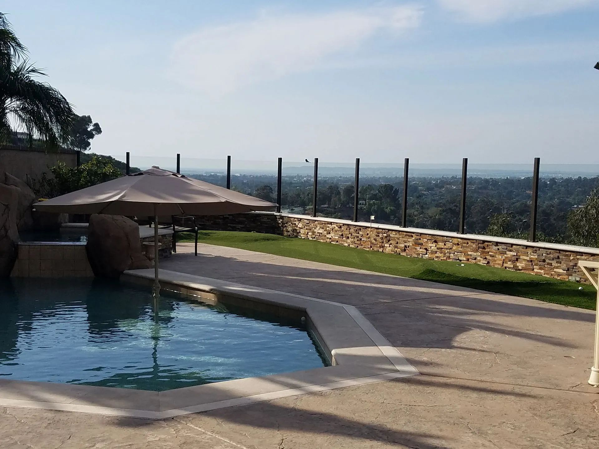 Backyard pool with a shaded lounge area, stone patio, and hillside city view beyond a glass fence