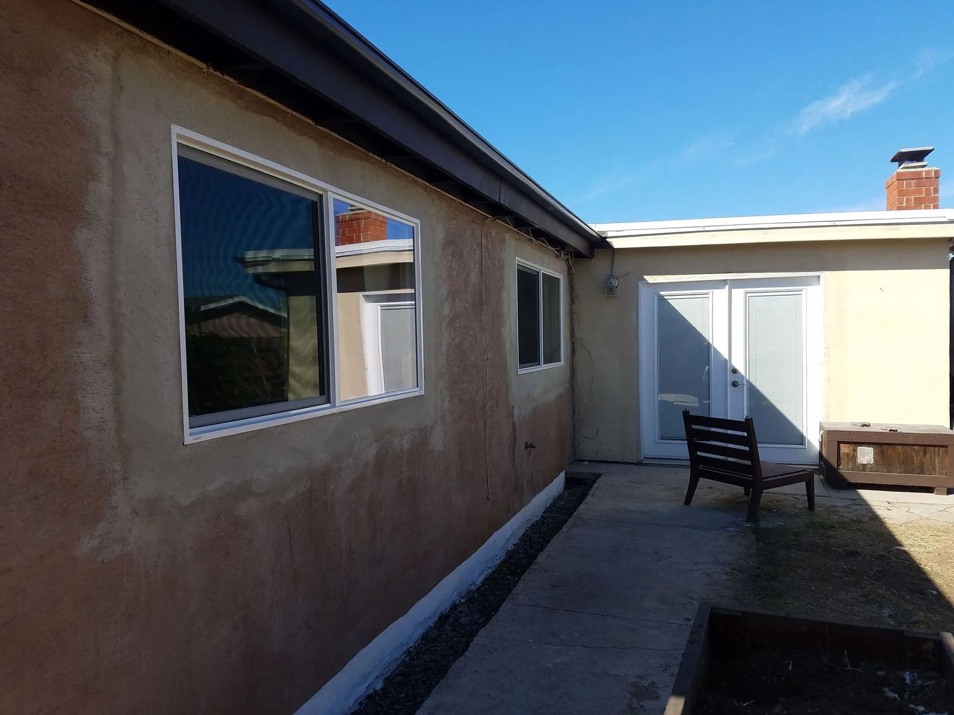 Narrow backyard patio between stucco walls with windows, a chair, and a white door under bright sunlight.